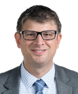 Headshot: Dark haired man in dark suit, white shirt and tie