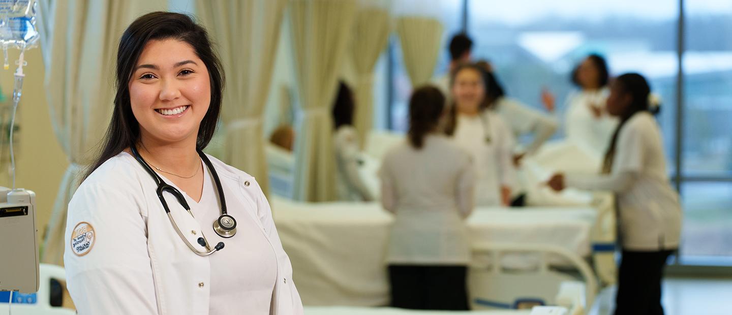 A nurse in a white lab coat, with a stethoscope around her neck, smiling at the camera.